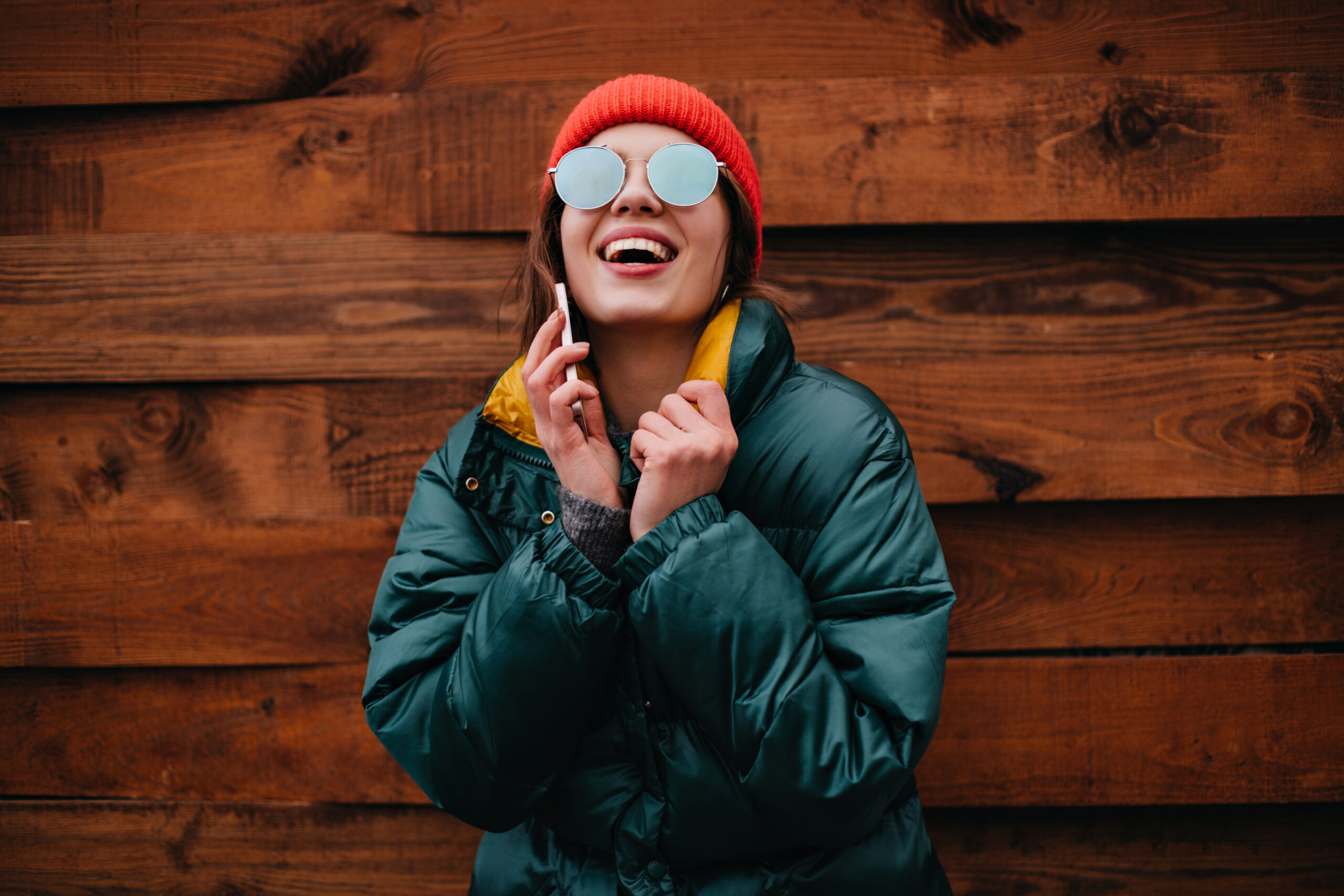 Young woman in great mood genuinely laughs while talking on phone. Girl in red hat and emerald color jacket poses on wooden background